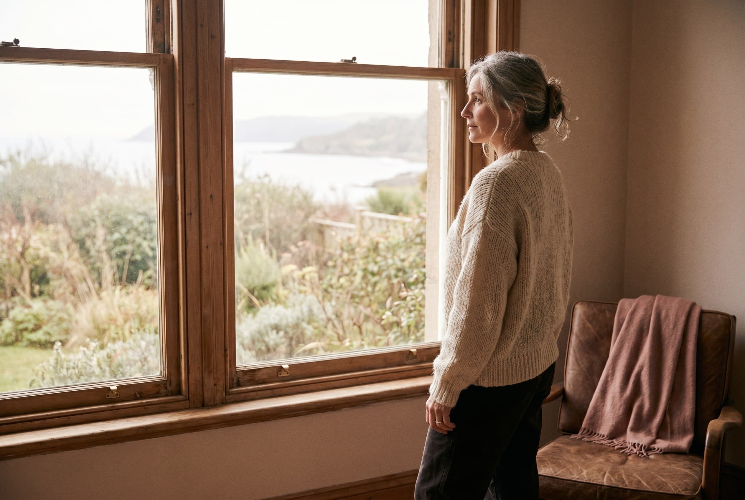 A woman in her 50s at a window looking out at a coastal view, in warm editorial light